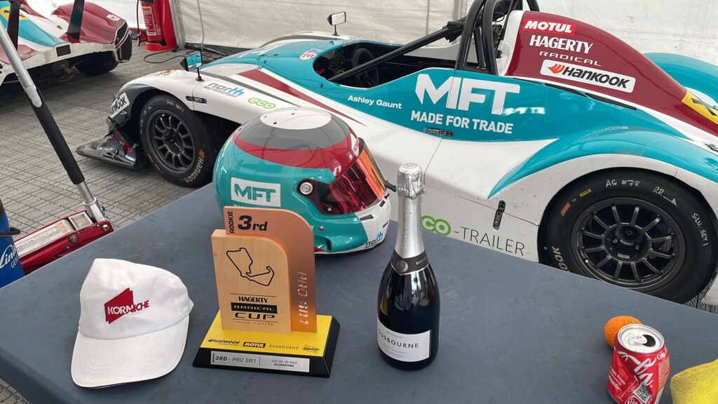 A Radical SR1 racing car with Made For Trade and Korniche branding parked in a Silverstone garage. Table displays racing helmet, hat, trophy, and champagne.