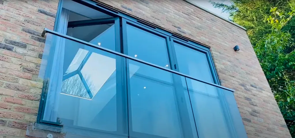 A modern open-plan living space with floor to ceiling bi-fold doors leading out to a glass balcony overlooking a garden.