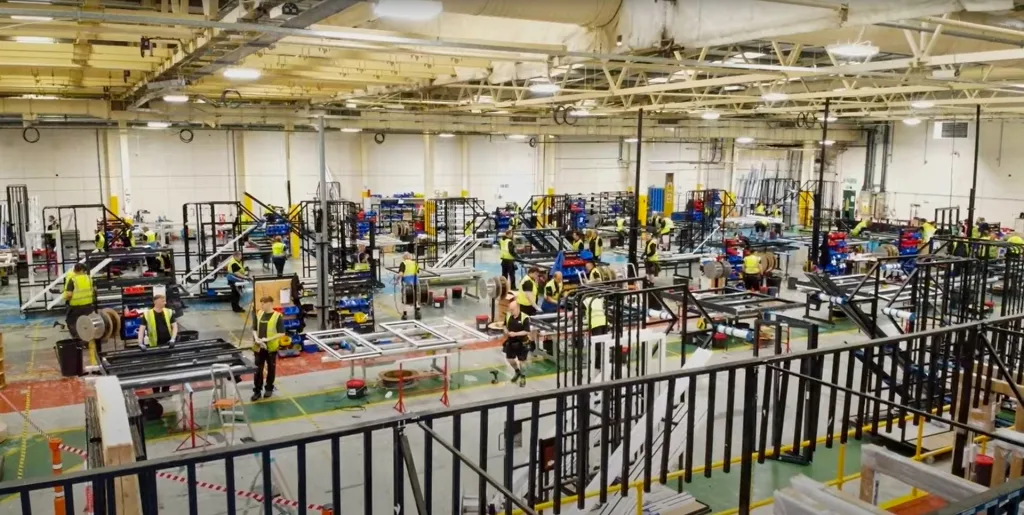 Photo of the Made for Trade factory interior showcasing the Korniche bi-fold door production line. The image shows several Korniche bi-fold door frames, at least five in a row, positioned along the production line. Made for Trade team members in high-visibility jackets are visible working on the doors.